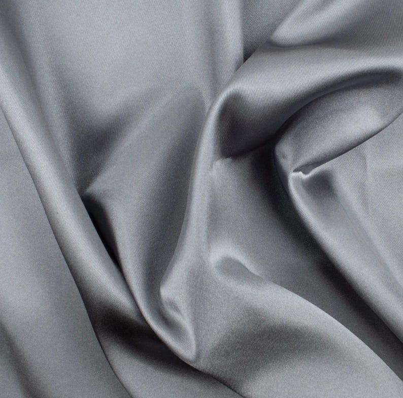 May include: A solid grey fabric with a smooth, lustrous finish. The material is draped in soft, flowing folds that create deep shadows and bright highlights across the surface, emphasizing the reflective quality and texture of the textile.