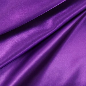 May include: A vibrant purple fabric with a smooth, lustrous finish and a reflective surface. The material is draped in soft, flowing folds that create deep shadows and bright highlights, emphasizing the silky texture and rich, saturated color.