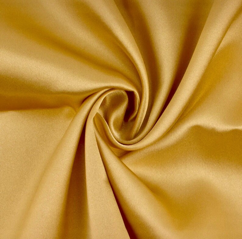 May include: A solid gold-colored fabric with a smooth, lustrous finish. The material is gathered into a central spiral, creating deep, flowing folds and shadows that highlight the reflective, satin-like texture of the textile surface.