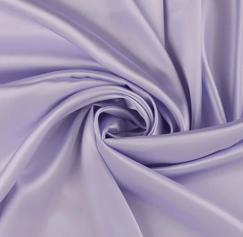 May include: A solid light lavender fabric with a smooth, lustrous finish. The material is draped in a central spiral pattern, creating deep, flowing folds and soft shadows that highlight the reflective, silky texture of the textile surface.