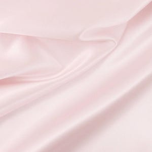 May include: A light pink fabric with a smooth, lustrous surface and a soft sheen. The material is draped in gentle, flowing folds that create subtle shadows and highlights across the textile, showcasing its reflective and silky texture.