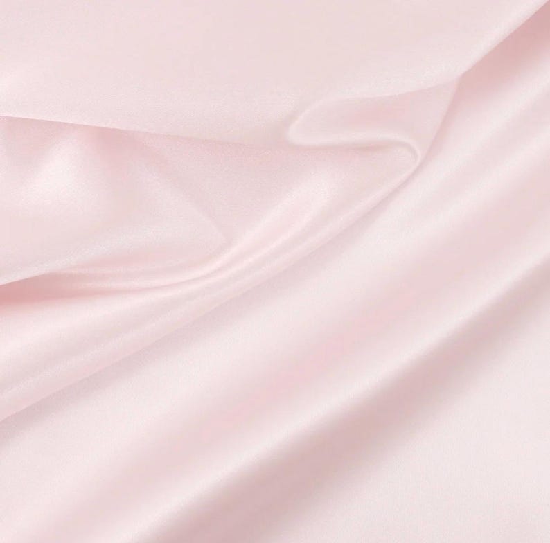 May include: A light pink fabric with a smooth, lustrous surface and a soft sheen. The material is draped in gentle, flowing folds that create subtle shadows and highlights across the textile, showcasing its reflective and silky texture.