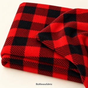 Peut inclure: Un tissu polaire plié présentant un motif à carreaux écossais rouge et noir vif. Le textile possède une surface douce et texturée avec un motif à carreaux répétitif. Le texte Bolthousefabric est imprimé en petites lettres noires en bas.