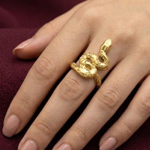 Könnte beinhalten: Ein goldfarbener Ring in Form einer gewundenen Schlange. Die Metalloberfläche weist ein strukturiertes, schuppenartiges Muster auf dem Körper der Schlange auf, die sich um den Finger windet, wobei der Kopf in Richtung des Fingerknöchels zeigt.