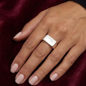 May include: A silver-toned signet ring featuring a smooth, rectangular flat top surface. The ring has a polished metallic finish and a solid, thick band design that sits horizontally across the finger, showcasing a clean and simple geometric shape.