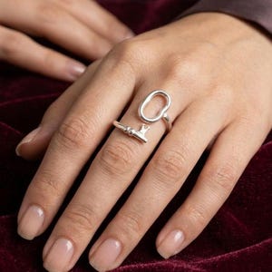 Pode incluir: Um anel de tom prateado com uma forma oval alongada e aberta que envolve o dedo. A banda termina num pequeno detalhe estilizado em forma de chave com uma extremidade entalhada, criando uma silhueta assimétrica distinta no dedo.