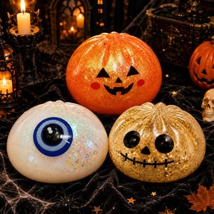Könnte beinhalten: Drei runde, glitzernde Halloween-Dekorationen. Eine ist ein orangefarbener Kürbis mit einem schwarzen Kürbisgesicht, eine ist ein weißer Augapfel mit einer großen blauen Iris und eine ist ein goldener Kürbis mit schwarzem Mund.