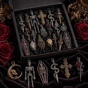May include: A collection of metal jewelry pendants featuring skeletons, coffins, skulls, snakes, crosses, and roses. The set includes various silver and gold-toned charms with intricate details, some accented with red enamel, displayed in a black box.