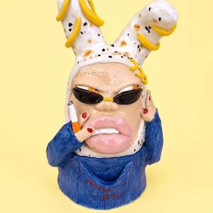 May include: A ceramic sculpture of a figure wearing a white bunny-eared hood with yellow accents and black spots. The figure wears black sunglasses, has large pink lips, and holds a cigarette. It is dressed in a blue denim-textured overall.