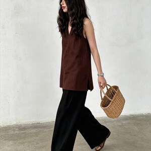 May include: A sleeveless dark brown tunic with a V-neckline and side slits, paired with loose-fitting black wide-leg trousers, brown open-toe sandals, and a small, light brown woven wicker basket with two top handles.
