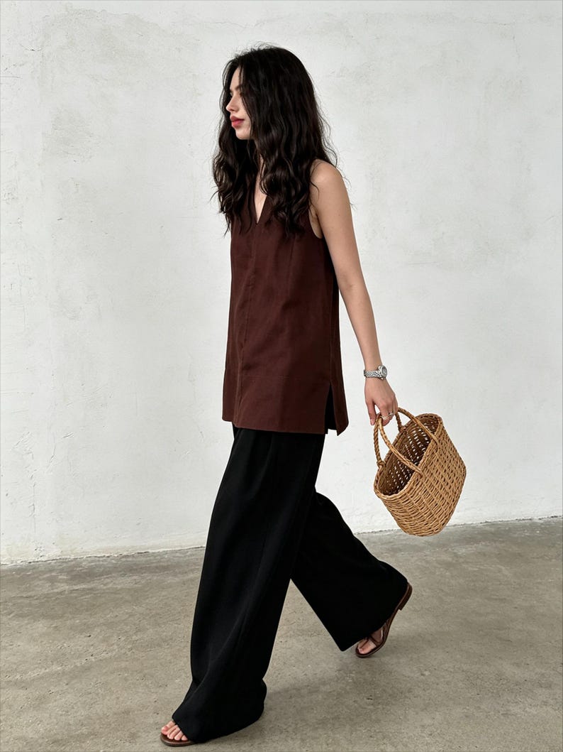 May include: A sleeveless dark brown tunic with a V-neckline and side slits, paired with loose-fitting black wide-leg trousers, brown open-toe sandals, and a small, light brown woven wicker basket with two top handles.