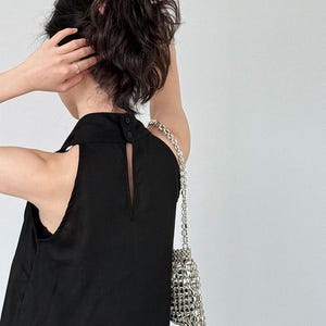 May include: A sleeveless black top featuring a high collar with two small black buttons at the back and a vertical keyhole opening. The garment is paired with a small, metallic silver handbag constructed from a grid of reflective, square-shaped beads.