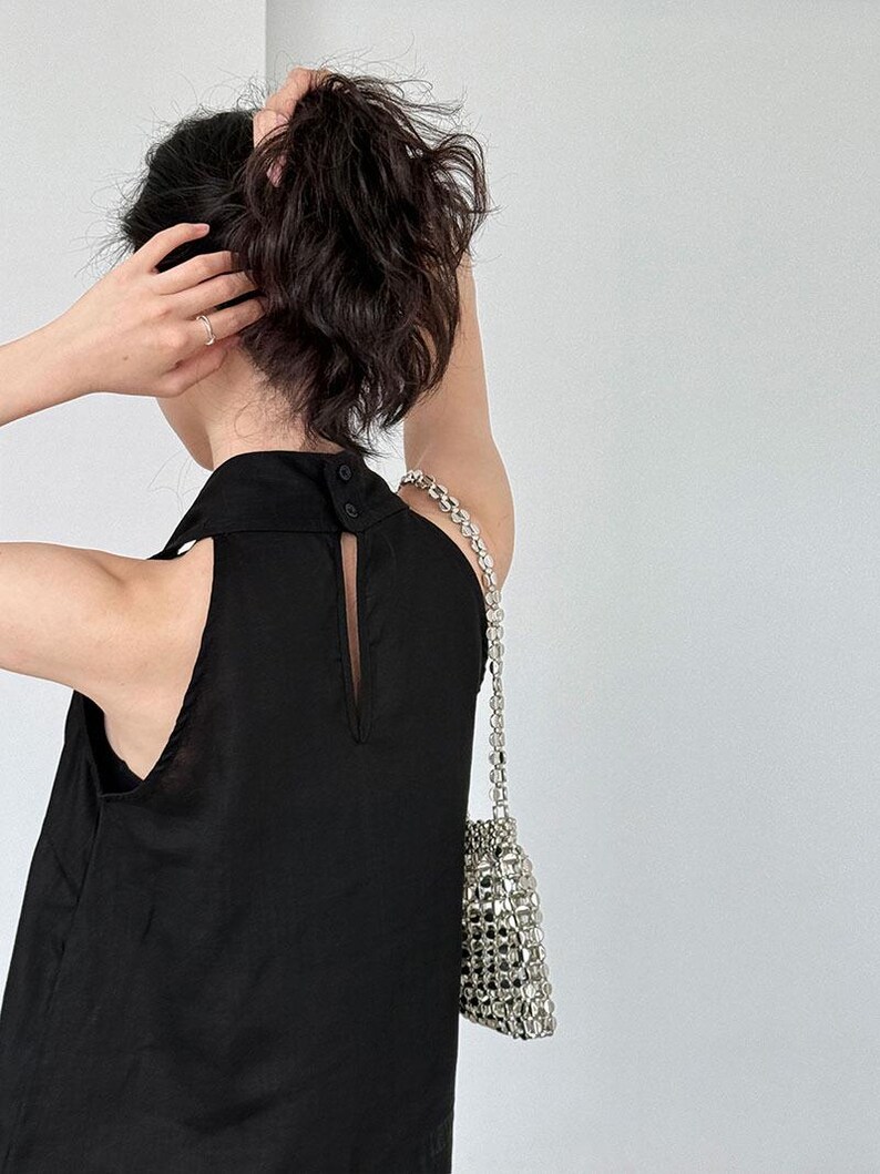 May include: A sleeveless black top featuring a high collar with two small black buttons at the back and a vertical keyhole opening. The garment is paired with a small, metallic silver handbag constructed from a grid of reflective, square-shaped beads.