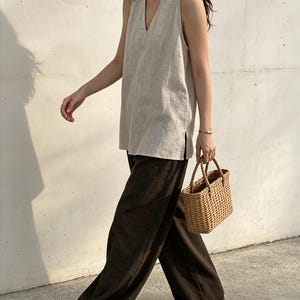 May include: A sleeveless beige V-neck top with side slits paired with dark brown wide-leg trousers and a small woven tan basket handbag. The outfit is completed with simple flat sandals, creating a neutral-toned ensemble with clean lines.
