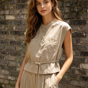 May include: A beige sleeveless button-down shirt with a mandarin collar, two chest pockets, and a thin matching cord tie at the waist. The shirt is paired with matching beige trousers featuring a pleated front design and side pockets.