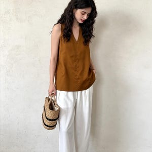 May include: A sleeveless brown V-neck top paired with loose-fitting white trousers featuring an elasticized drawstring waist. The outfit is accessorized with a woven straw handbag with black horizontal stripes and simple open-toe sandals.