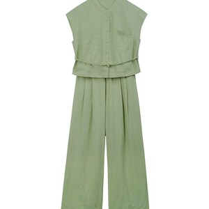 May include: A light sage green sleeveless jumpsuit featuring a mandarin collar, a button-down front closure, two chest pockets, and a thin matching fabric belt at the waist. The garment has wide-leg trousers with subtle pleats at the front.