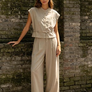 May include: A beige two-piece set consisting of a sleeveless button-down top with a mandarin collar and two chest pockets, paired with matching high-waisted wide-leg trousers featuring a thin tie-waist detail and a pleated front design.