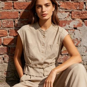 May include: A beige sleeveless button-down shirt with a mandarin collar, two chest pockets, and a thin drawstring waist tie. The shirt is paired with matching beige trousers, creating a coordinated set with a structured, utility-inspired design.