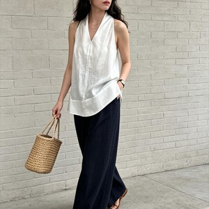 May include: A sleeveless white V-neck top paired with loose-fitting black wide-leg trousers. The outfit is accessorized with a woven straw bucket bag and brown leather thong sandals, creating a simple and functional ensemble for warm weather.