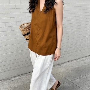 May include: A sleeveless brown top with a V-neckline paired with loose-fitting white trousers. The outfit is accessorized with a woven straw tote bag featuring black trim and brown slip-on loafers, creating a coordinated and neutral ensemble.
