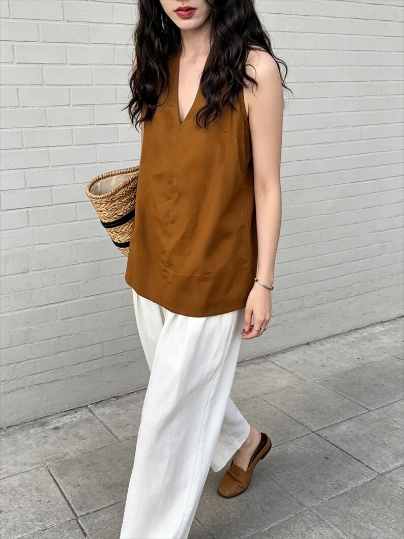 May include: A sleeveless brown top with a V-neckline paired with loose-fitting white trousers. The outfit is accessorized with a woven straw tote bag featuring black trim and brown slip-on loafers, creating a coordinated and neutral ensemble.