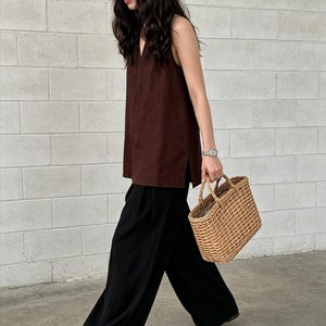 May include: A sleeveless dark brown V-neck top with side slits paired with loose-fitting black wide-leg trousers and a rectangular woven straw tote bag with two top handles, worn with simple brown flat sandals.