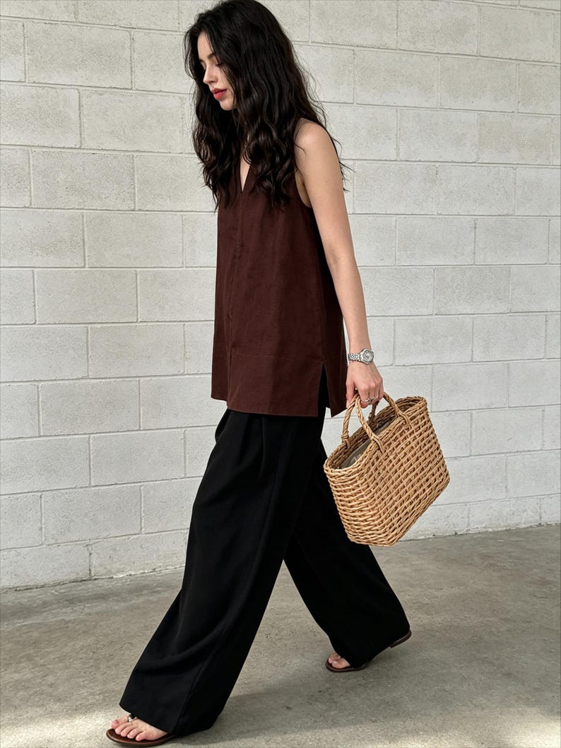 May include: A sleeveless dark brown V-neck top with side slits paired with loose-fitting black wide-leg trousers and a rectangular woven straw tote bag with two top handles, worn with simple brown flat sandals.