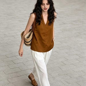May include: A sleeveless brown V-neck top paired with loose-fitting white trousers, a woven straw tote bag with black horizontal stripes, and brown leather loafers. The ensemble features a clean, simple aesthetic with neutral earth tones.