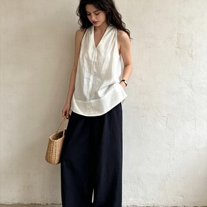 May include: A sleeveless white V-neck top with a textured finish paired with loose-fitting black wide-leg trousers. The outfit is accessorized with a woven straw handbag and tan slip-on shoes, creating a coordinated ensemble of neutral tones.