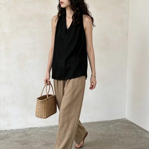 May include: A sleeveless black top with a V-neckline paired with loose-fitting tan trousers. The outfit is accessorized with a small, woven straw basket bag featuring two top handles and simple brown thong sandals on the feet.
