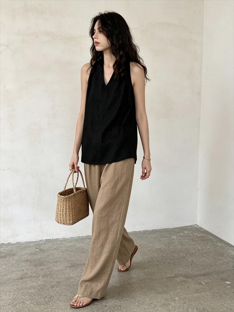 May include: A sleeveless black top with a V-neckline paired with loose-fitting tan trousers. The outfit is accessorized with a small, woven straw basket bag featuring two top handles and simple brown thong sandals on the feet.