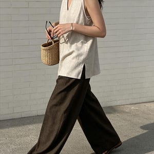 May include: A sleeveless beige V-neck top with side slits paired with dark brown wide-leg trousers. The outfit includes a small woven straw basket bag, black-rimmed glasses, a thin silver bracelet, and brown open-toe sandals.