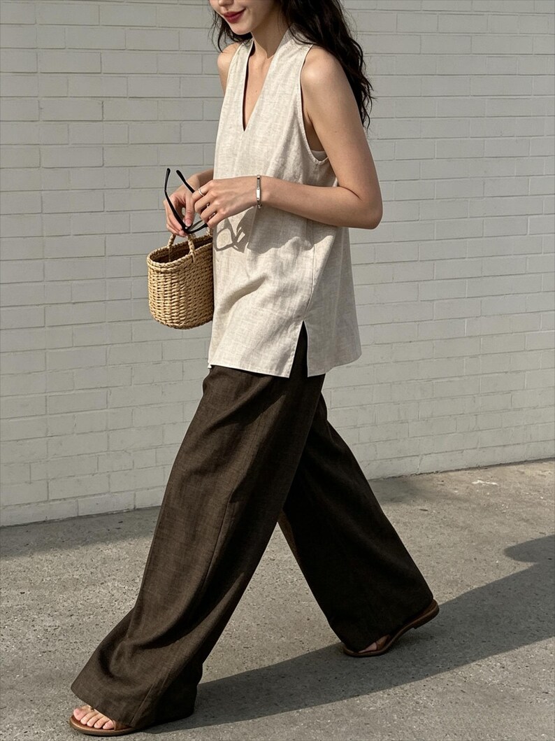 May include: A sleeveless beige V-neck top with side slits paired with dark brown wide-leg trousers. The outfit includes a small woven straw basket bag, black-rimmed glasses, a thin silver bracelet, and brown open-toe sandals.