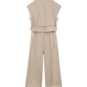 May include: A beige sleeveless jumpsuit featuring a mandarin collar, a button-down front closure, and two chest pockets. The garment includes a thin, integrated waist tie and wide-leg trousers, presenting a clean, monochromatic aesthetic.
