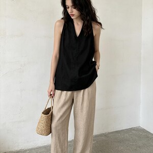May include: A sleeveless black top with a V-neckline paired with loose-fitting beige trousers and a woven straw handbag. The outfit features a simple design with clean lines, showcasing a neutral color palette and a relaxed, comfortable fit.