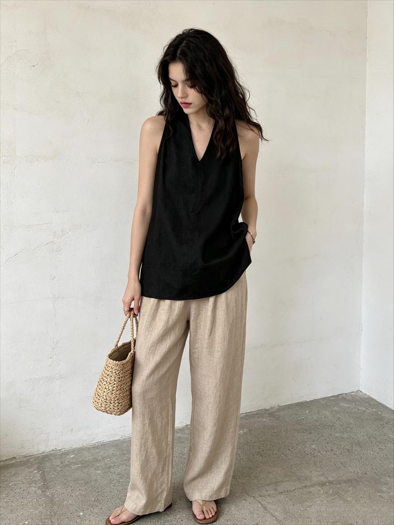 May include: A sleeveless black top with a V-neckline paired with loose-fitting beige trousers and a woven straw handbag. The outfit features a simple design with clean lines, showcasing a neutral color palette and a relaxed, comfortable fit.