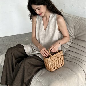 May include: A sleeveless beige linen top with a V-neckline paired with wide-leg dark brown trousers and a small rectangular woven wicker basket bag with two top handles. The outfit is composed of neutral-toned garments and a natural fiber accessory.