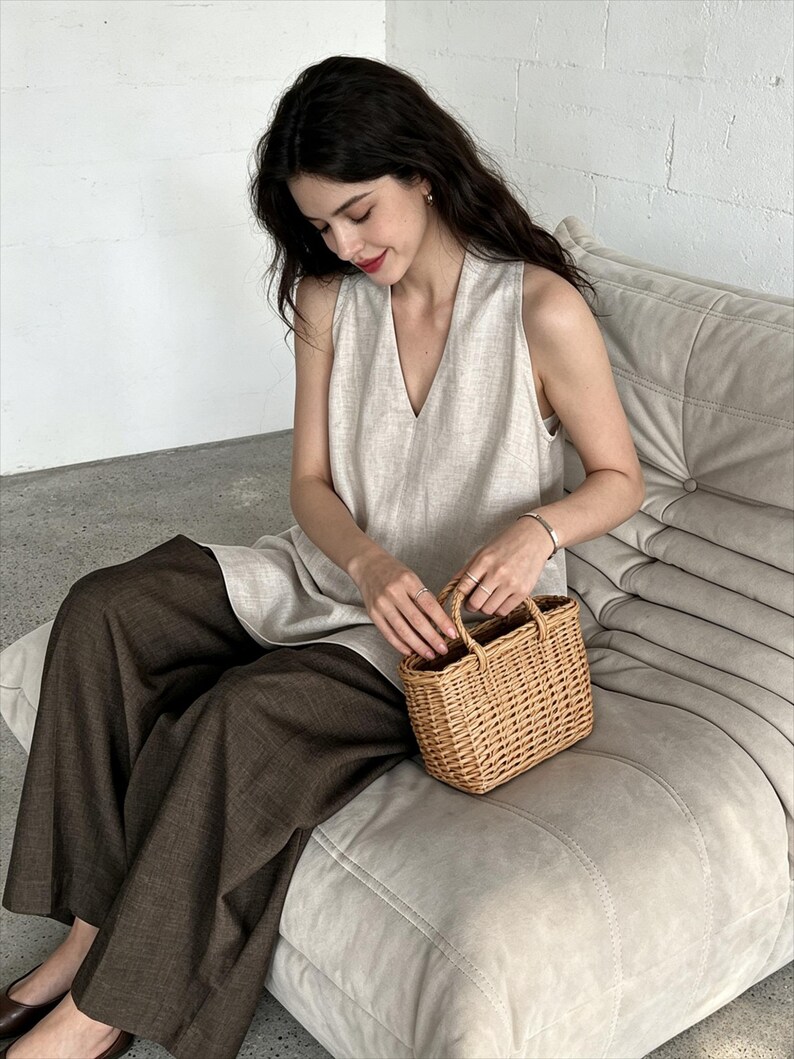 May include: A sleeveless beige linen top with a V-neckline paired with wide-leg dark brown trousers and a small rectangular woven wicker basket bag with two top handles. The outfit is composed of neutral-toned garments and a natural fiber accessory.