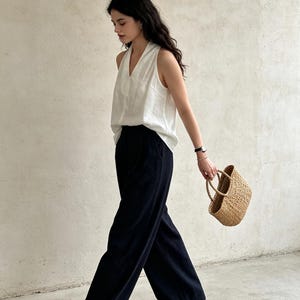 May include: A sleeveless white V-neck top paired with high-waisted black wide-leg trousers. The outfit is accessorized with a small woven straw handbag, a metallic wristwatch, and tan flat shoes, creating a clean and coordinated ensemble.