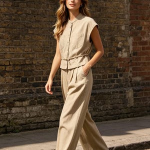 May include: A beige sleeveless button-down shirt with a mandarin collar and two chest pockets, paired with matching high-waisted wide-leg trousers featuring a thin tie-waist belt. The outfit is a solid neutral tan color with visible seam detailing.