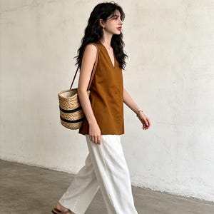 May include: A sleeveless brown V-neck top paired with wide-leg white trousers, a woven straw bucket bag with black horizontal stripes, and tan flat shoes. The outfit features a clean, simple silhouette with neutral earth tones and textures.