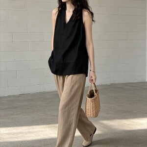 May include: A black sleeveless V-neck top paired with wide-leg tan trousers and beige flat shoes. The outfit is accessorized with a small, light brown woven wicker handbag featuring two top handles, creating a coordinated ensemble.