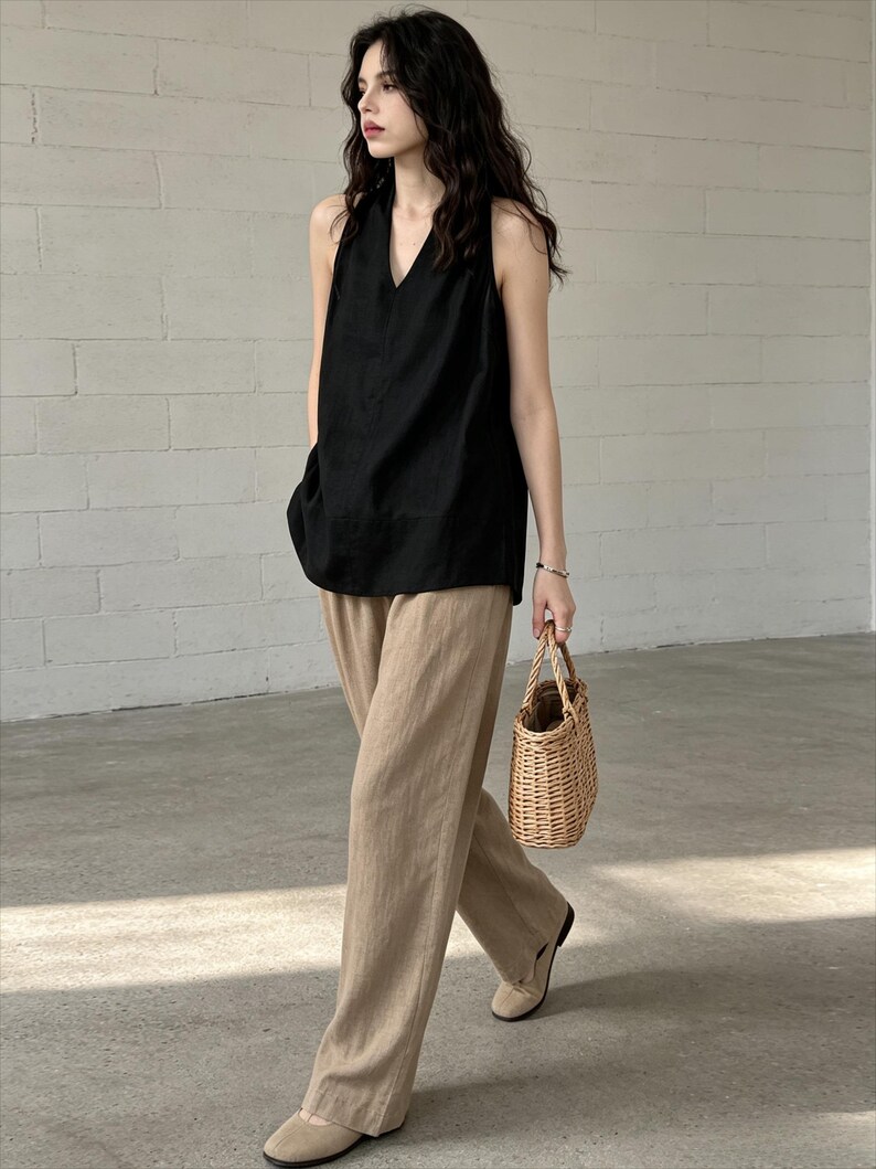 May include: A black sleeveless V-neck top paired with wide-leg tan trousers and beige flat shoes. The outfit is accessorized with a small, light brown woven wicker handbag featuring two top handles, creating a coordinated ensemble.