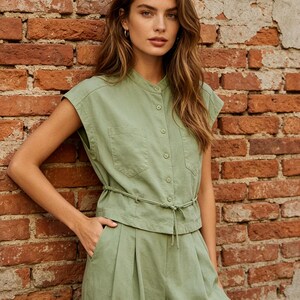 May include: A sage green sleeveless button-down shirt with a mandarin collar, two chest pockets, and a thin drawstring waist tie, paired with matching high-waisted pleated trousers that feature side pockets and a relaxed, straight-leg silhouette.