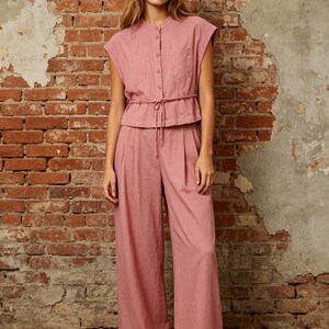 May include: A dusty rose two-piece set consisting of a sleeveless button-down top with a mandarin collar, two chest pockets, and a drawstring waist, paired with matching high-waisted wide-leg trousers featuring front pleats and a relaxed fit.
