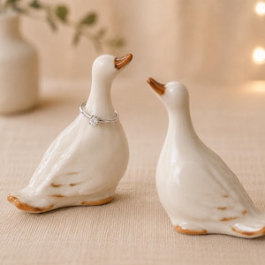 Może przedstawiać: Dwie białe ceramiczne figurki kaczek z brązowymi akcentami na dziobach i ogonach. Kaczka po lewej stronie ma na szyi cienki srebrny pierścionek z małym przezroczystym kamieniem. Figurki kaczek są gładkie, błyszczące i przedstawione w pozycji siedzącej.