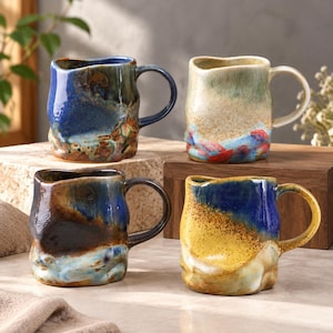 May include: Four ceramic mugs with irregular, organic shapes and glossy finishes. One is deep blue with brown accents, one is cream with red and blue details, one is dark brown and blue, and one is yellow and blue with a speckled texture.