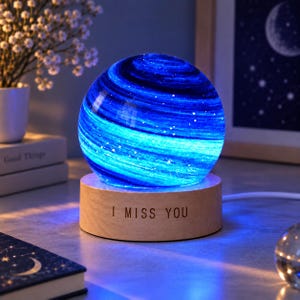 Puede incluir: Una lámpara esférica con un patrón de nebulosa azul y blanco que brilla con luz interna. La esfera descansa sobre una base cilíndrica de madera clara con el texto I MISS YOU grabado en negro en la superficie frontal.