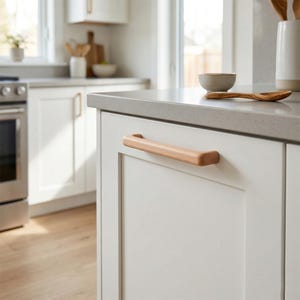 May include: A light-coloured wooden cabinet handle with a smooth, rounded rectangular shape attached to a white kitchen cabinet door. The handle features a natural wood grain finish and is mounted horizontally on the flat panel of the cabinet door.